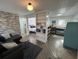 A compact living space with a sofa, a small table, a built-in bookshelf, a kitchenette, and a bathroom with a shower.