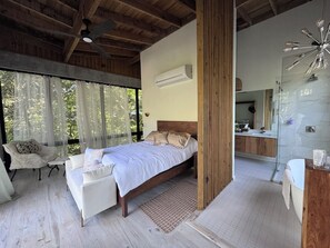 Ein Schlafzimmer mit Bett, Sessel, großem Fenster und einem Badezimmer mit Spiegel und Waschbecken.