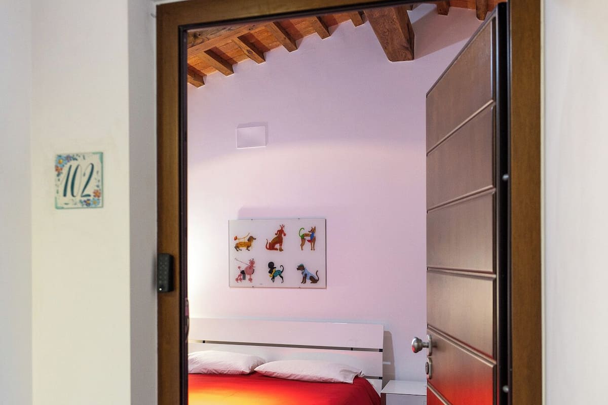 Quarto com colcha vermelha, mesa de cabeceira branca e uma obra de arte na parede.