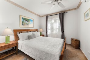 Ein Schlafzimmer mit einem Bett, zwei Nachttischen mit Lampen, einem Deckenventilator, einem Fenster mit Vorhängen und einem gerahmten Bild an der Wand.