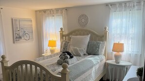 Ein Schlafzimmer mit einem Bett, zwei Nachttischen, einer Lampe, einem Wanddekor und einem Fenster mit Vorhängen.