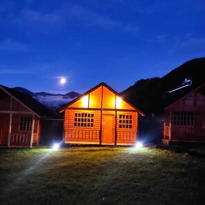 Eine Holzhütte mit einer mittleren Tür und Fenstern, umgeben von einer grünen Wiese und Bergen im Hintergrund.