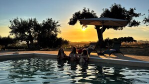 A swimming pool with three people relaxing, surrounded by trees and a sunset view.