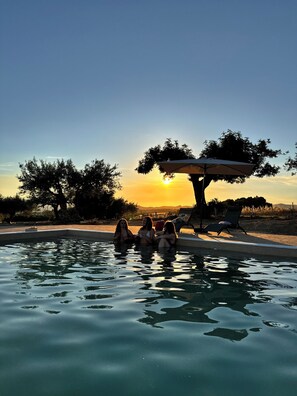 A swimming pool with three people relaxing, surrounded by trees and a sunset view.