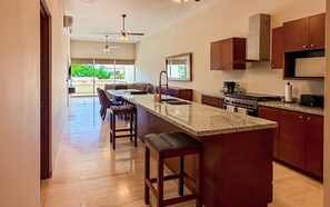 A modern kitchen with a central island, bar stools, a dining area with a sofa, and a view of greenery outside. - Private Terrace with Views, Pool & Gym Access (Cabo San Lucas)