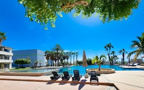 Uma área de piscina com espreguiçadeiras, palmeiras e um edifício moderno ao fundo.