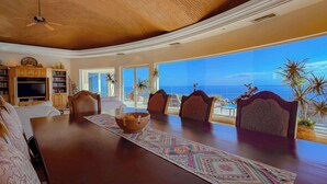 Un salón espacioso con una gran mesa de comedor de madera, asientos cómodos, televisión y vista al océano.
