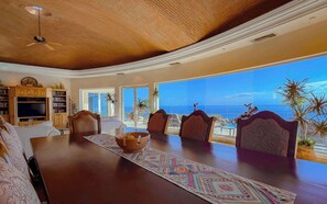 Ein großzügiges Wohnzimmer mit einem großen Holzesstisch, bequemen Sitzmöglichkeiten, einem Fernseher und Blick auf den Ozean.