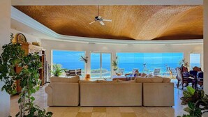 Un salón amplio con un sofá grande, un ventilador de techo y vistas al océano a través de amplios ventanales.