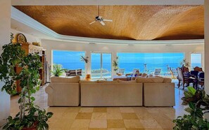 Ein geräumiges Wohnzimmer mit einer großen Couch, einem Deckenventilator und Meerblick durch große Fenster.