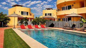 A swimming pool with red lounge chairs, surrounded by a wooden fence and yellow buildings. - Ground Floor Condo with Pool, Near Cerritos Beach (El Pescadero)