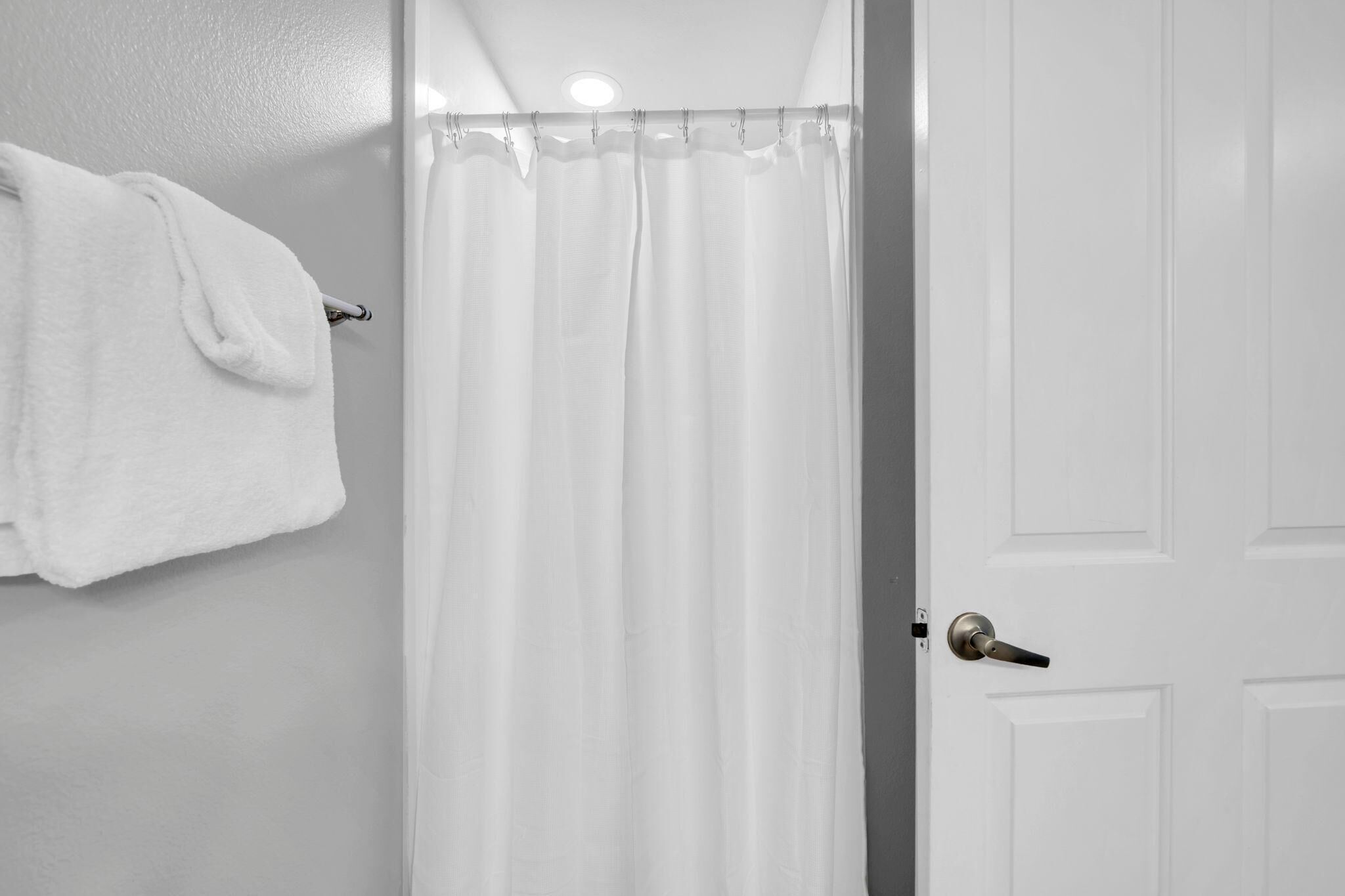 Un baño con cortina de ducha blanca, una toalla blanca colgada en un perchero y una puerta blanca con pomo plateado.