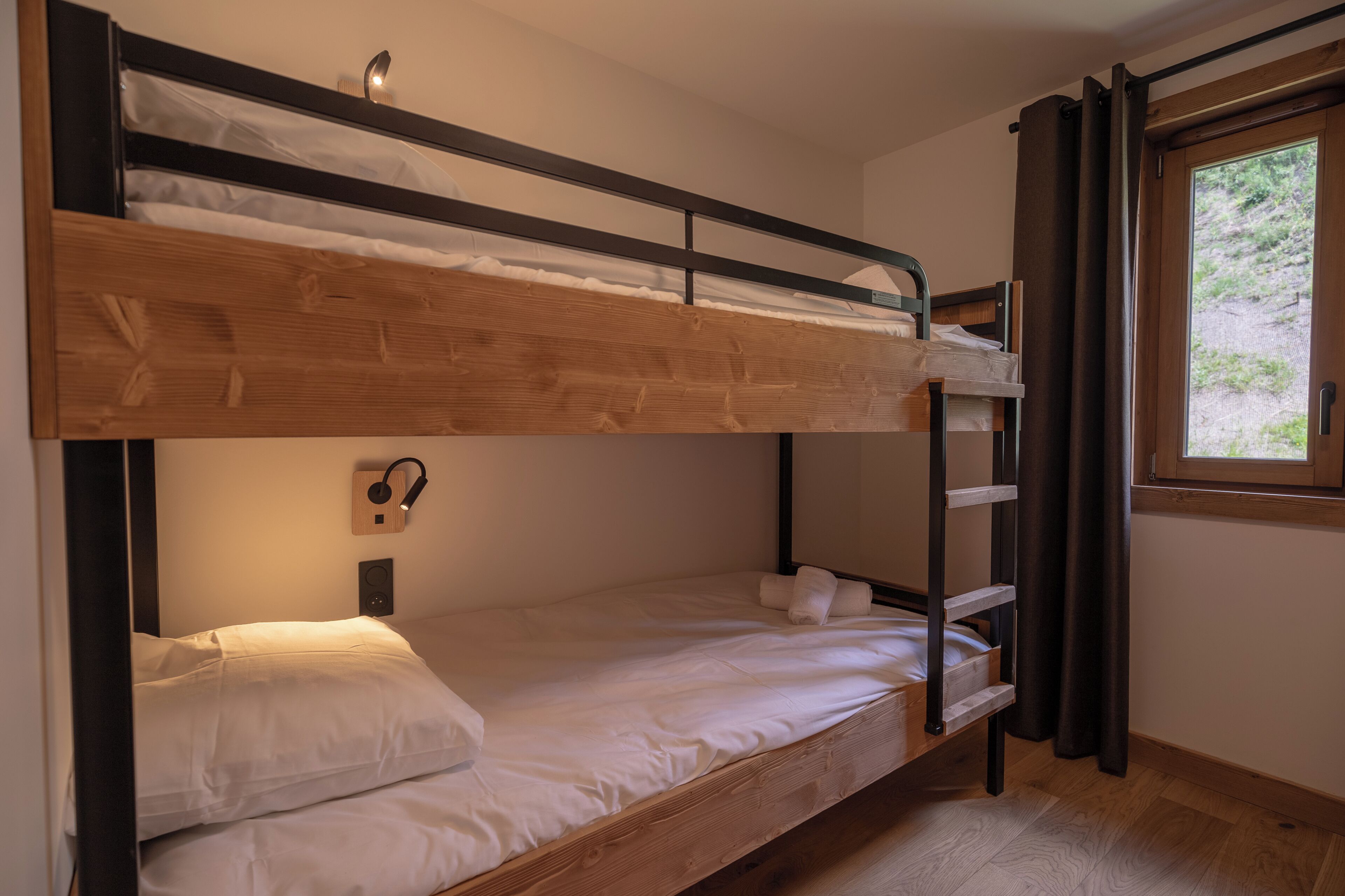 A bunk bed with a wooden frame, a wall-mounted light, and a window with curtains.