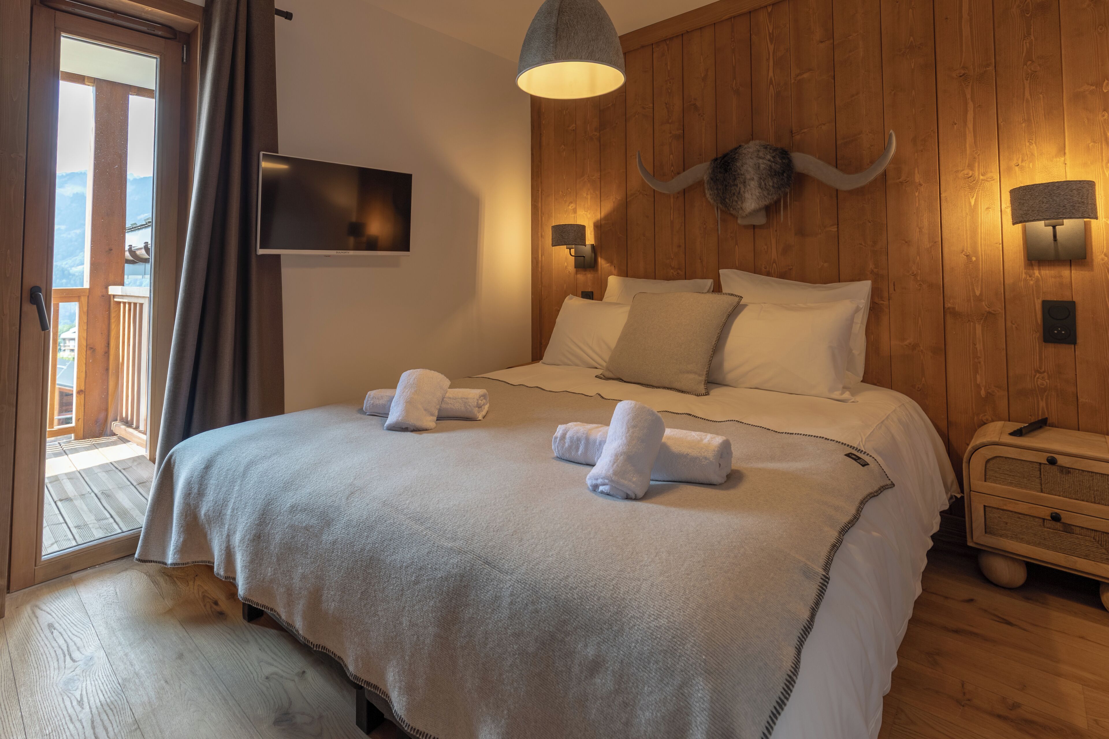 Habitación de hotel con cama, televisor montado, pared de madera con decoración de cabeza de toro y mesita de noche de madera.