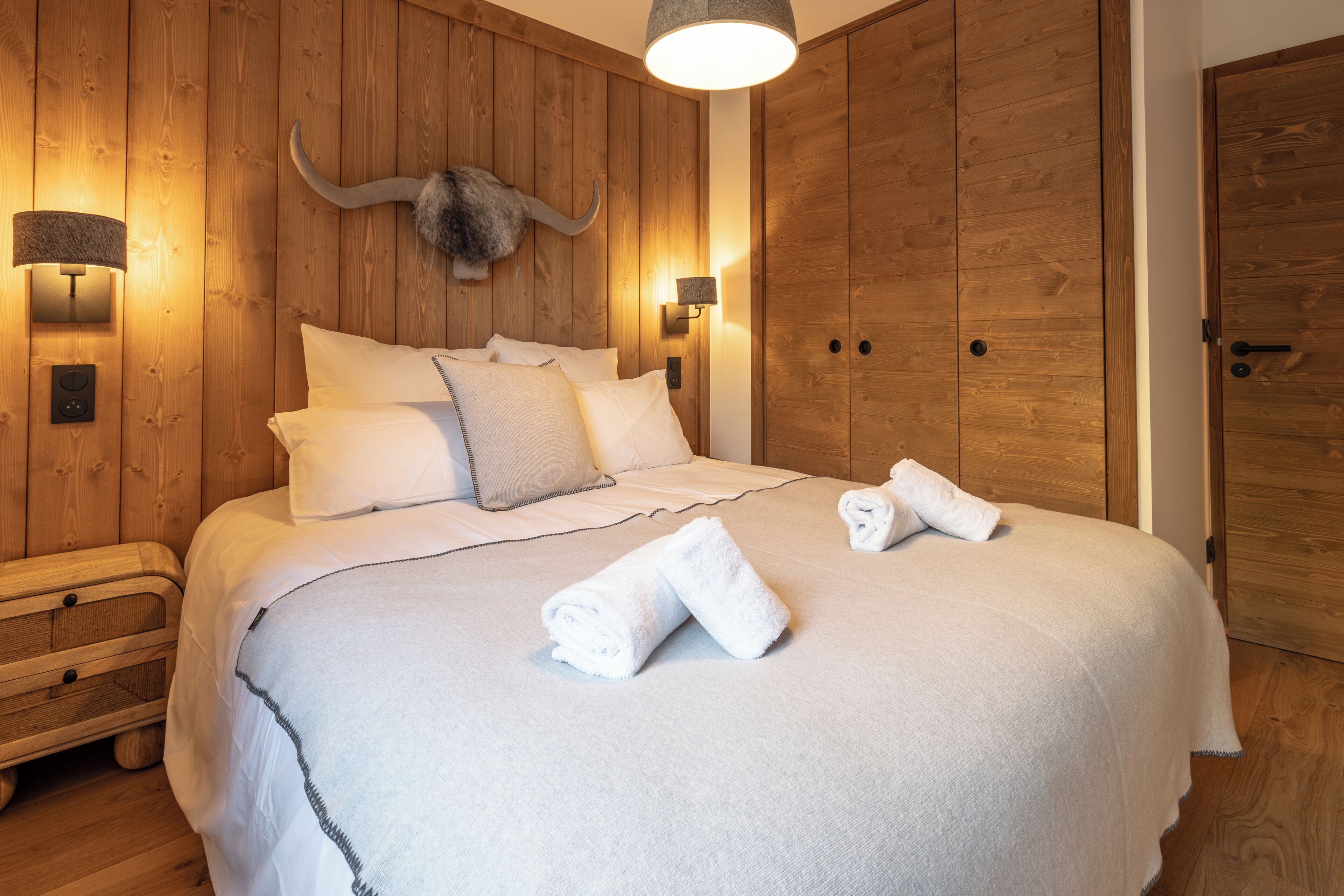 Une pièce avec un mur en bois, un lit avec une literie blanche, deux lampes fixées au mur et une grande porte en bois.