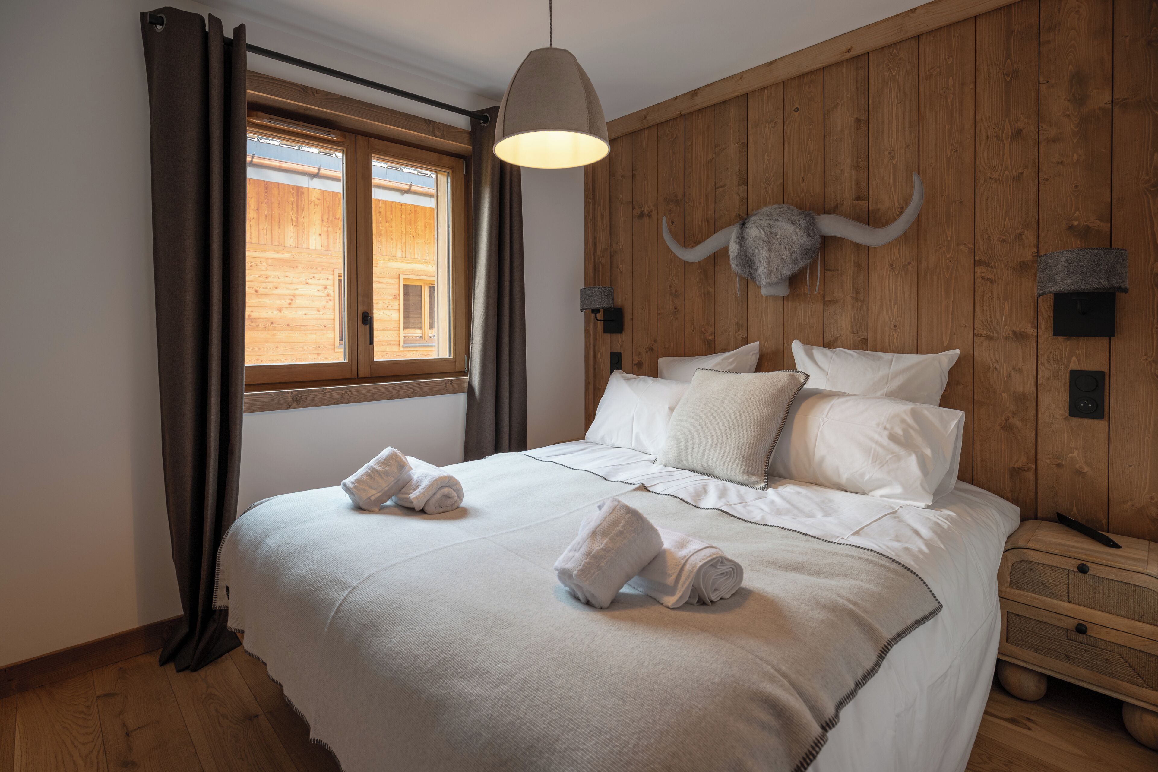 Un dormitorio con cabecera de madera, una cama grande con ropa blanca, una lámpara colgante y una ventana con cortinas.