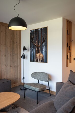 A modern living room with a wooden wall, a framed deer picture, a round wooden table, a grey upholstered chair, and a black floor lamp. - TERRESENS - Les Balcons de Juliette (Notre-Dame-de-Bellecombe)