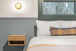 En seng med hvid og ternet sengetøj, et natbord med skuffe, en vægmonteret lampe og et vindue med udsigt til træer.