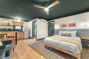 Quarto de hotel moderno com cama, escrivaninha, cadeira, ventilador de teto e cozinha compacta.