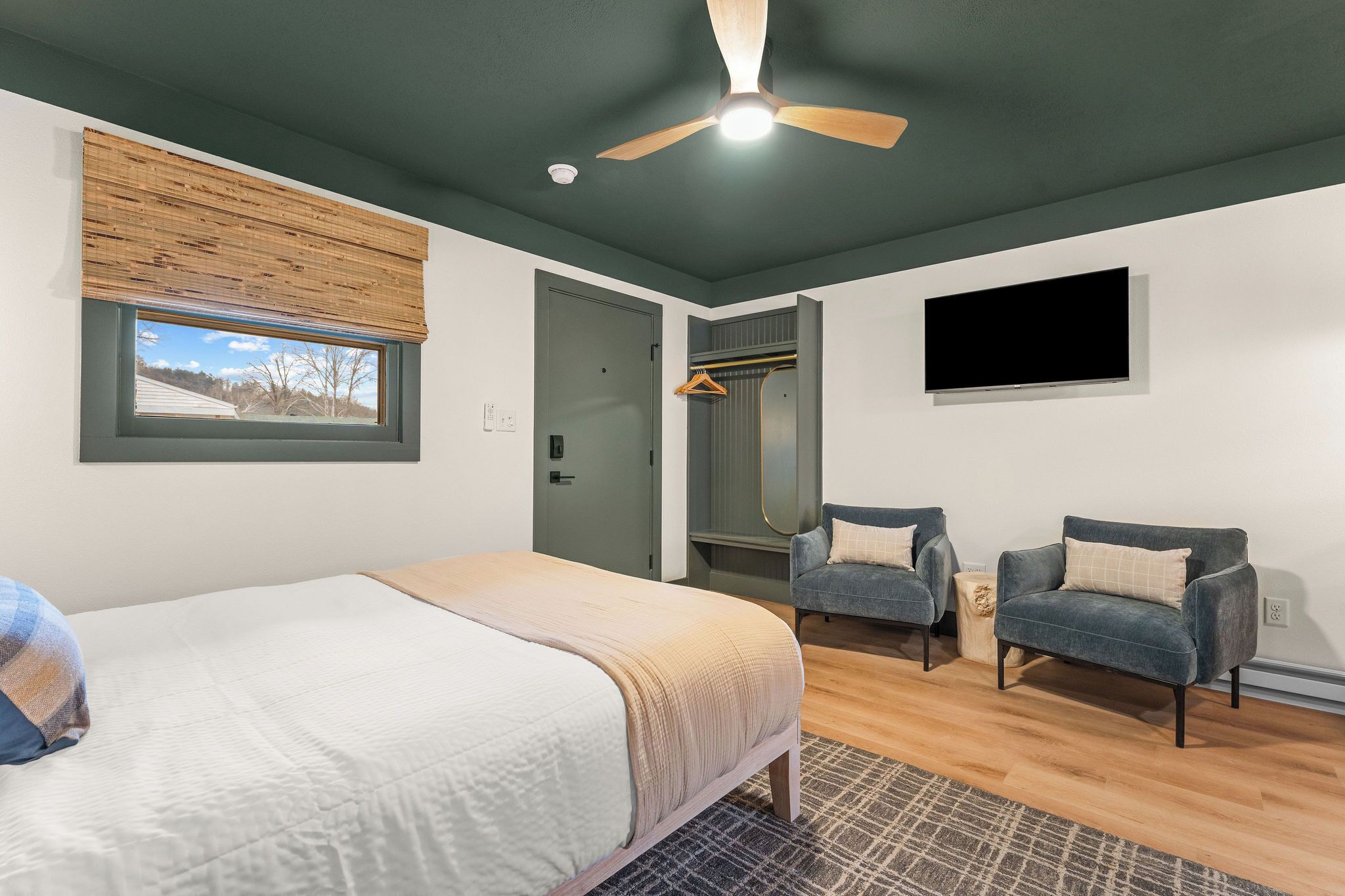 A bedroom with a bed, two armchairs, a ceiling fan, a TV, and a window with blinds.
