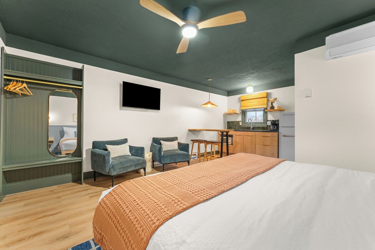A bedroom with a bed, two chairs, a small table, a ceiling fan, a TV, and a kitchenette.