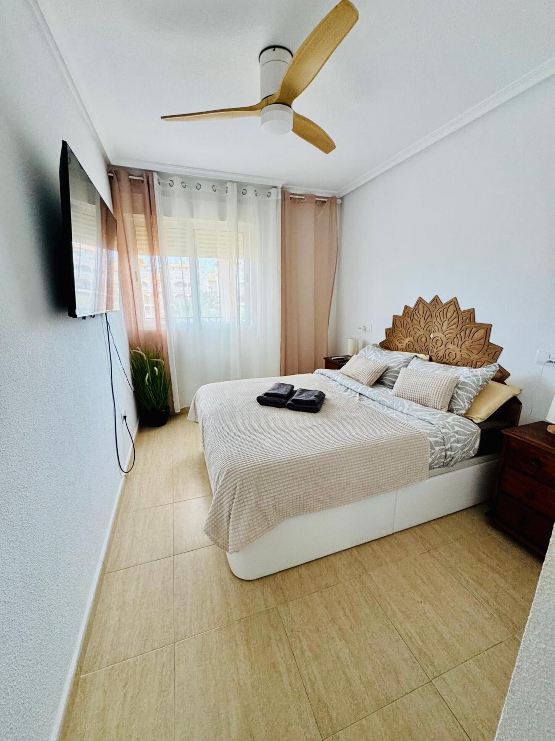 Un dormitorio con cama, ventilador de techo, televisor montado en la pared y ventana con cortinas.