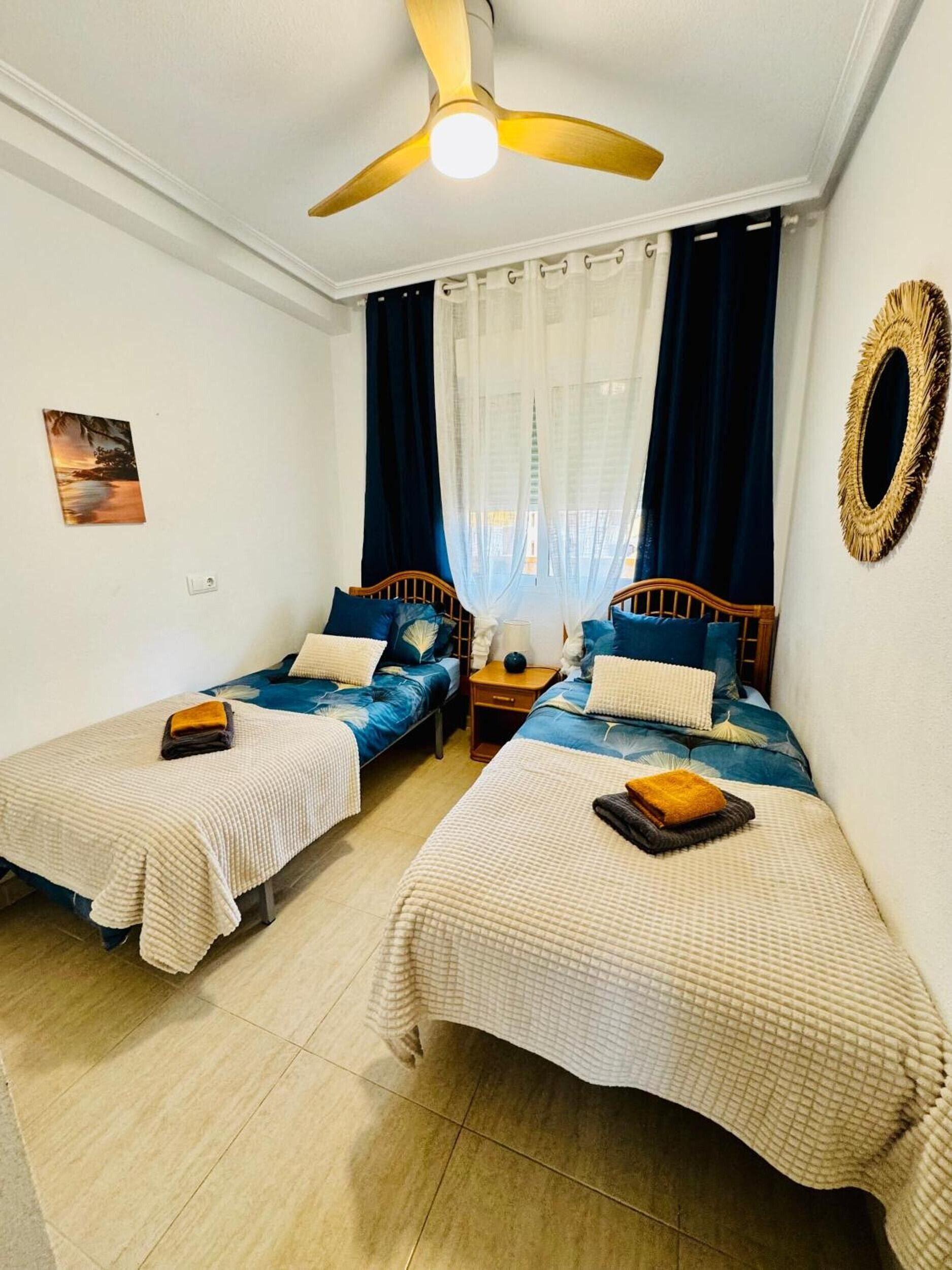 Habitación con dos camas, ventilador de techo, ventana con cortinas y espejo montado en la pared.