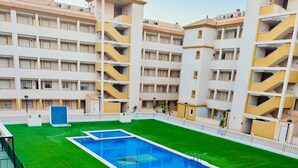 A rooftop pool surrounded by a green lawn, with a white fence and a building with multiple balconies in the background.