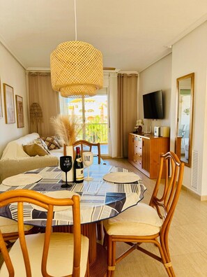 Uma área de refeições com uma mesa redonda, cadeiras, uma luminária pendente trançada, uma televisão e uma varanda com vista.