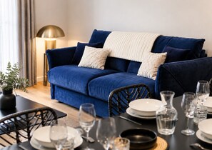 A blue sofa with white and dark blue cushions, a table set with plates and glasses, and a floor lamp. - Outremer – Blue Comfort in the Heart of Colmar (Colmar)