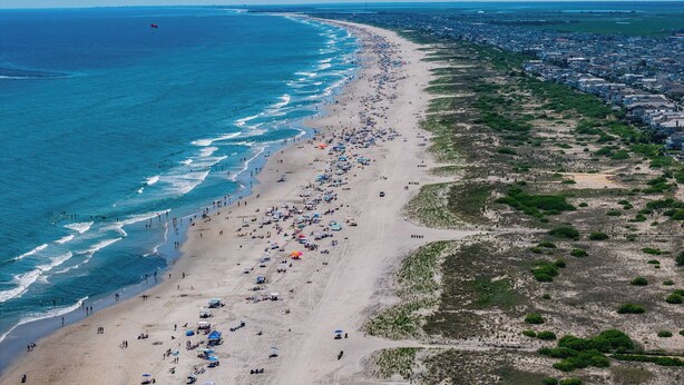 An aerial view of a beach with a long stretch of sand, a few umbrellas, and a distant cityscape. - 2 Blocks to Beach & Boardwalk – 3BR/2BA in Gold Coast, OCNJ (Ocean City)