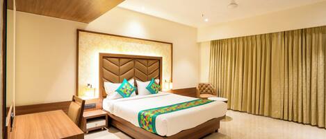 A hotel room with a large bed, a wooden headboard, a bedside table, a nightstand, a lamp, and a window with curtains.