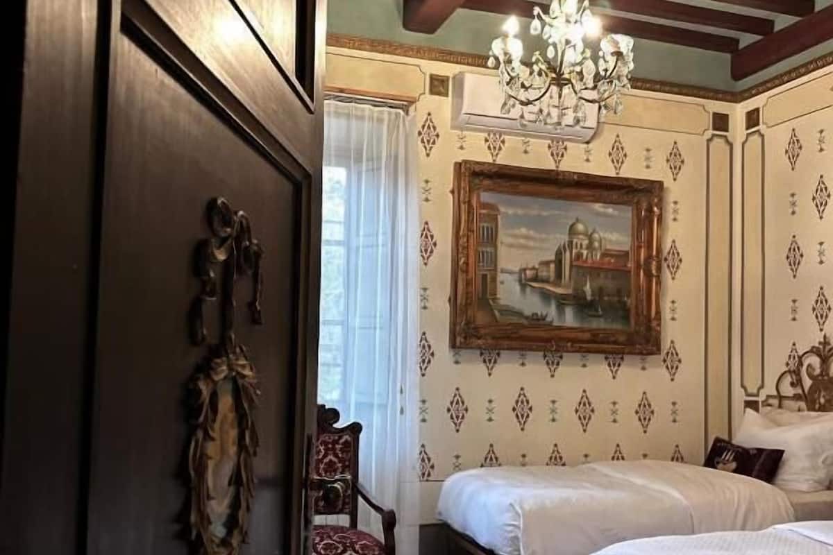 A room with two beds, a chandelier, a painting on the wall, and a patterned carpet.