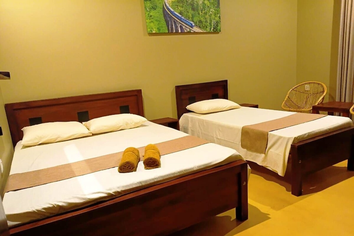 Two wooden beds with white linens and towels, a wicker chair, and a painting of a bridge on the wall.