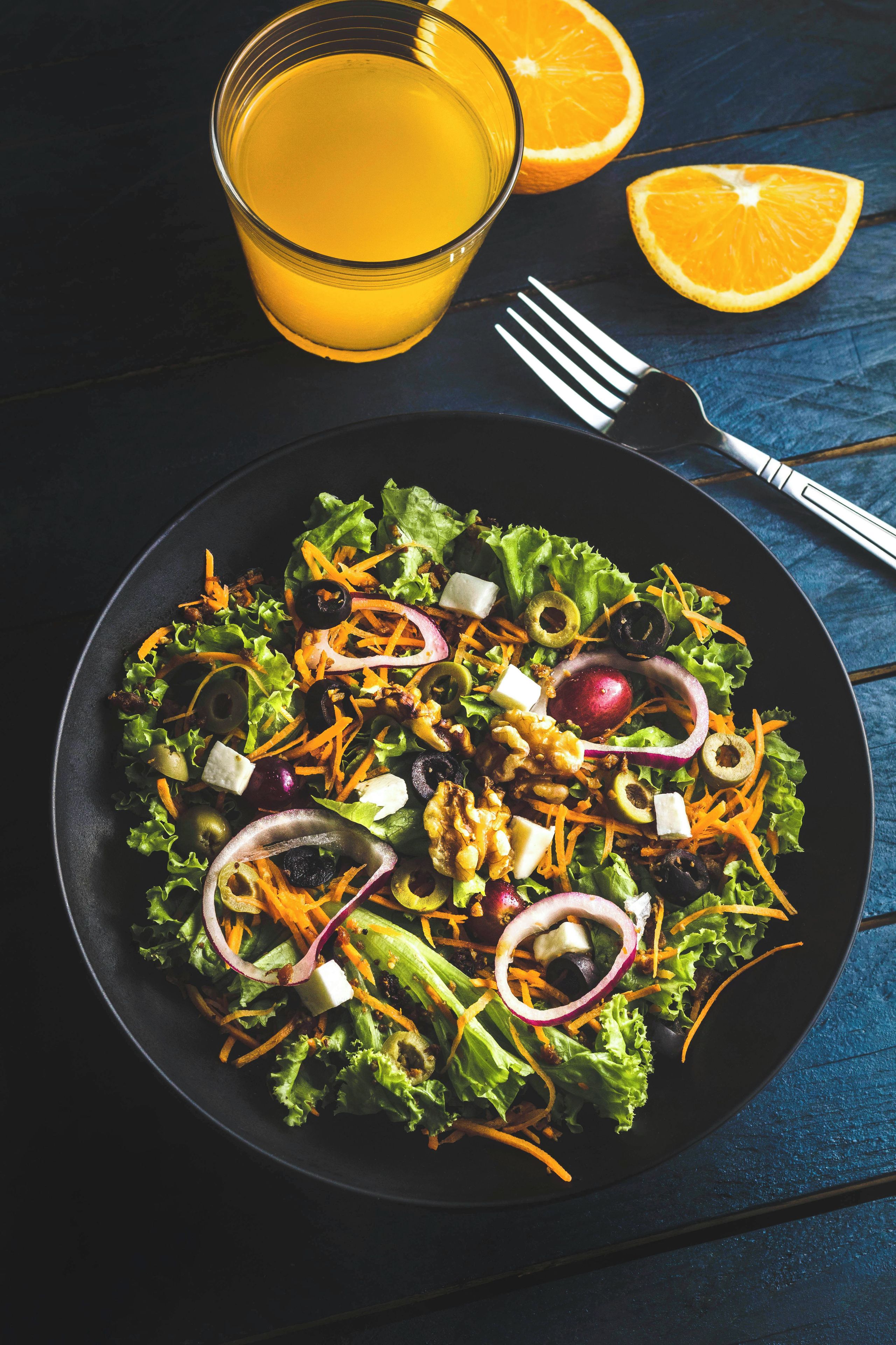 Un piatto di insalata con diverse verdure e noci, un bicchiere di succo d'arancia e una forchetta su un tavolo scuro.