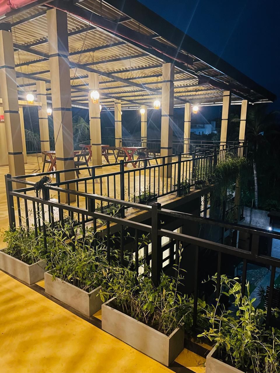 Un espace extérieur abrité, doté d’une rambarde en métal, de jardinières en béton agrémentées de plantes et d’une série de luminaires.