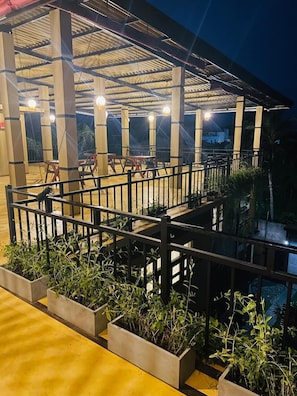 Un espacio exterior techado con barandilla metálica, macetas de concreto con plantas verdes y una serie de luces.