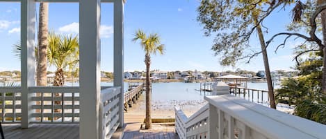 Una terraza de madera con baranda blanca que da a un puerto deportivo con barcos y una playa.