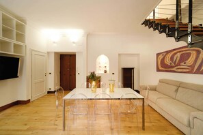 Ein Essbereich mit Tisch, Stühlen und einer Couch. An der Wand ist ein Fernseher angebracht und über der Couch hängt ein Gemälde.