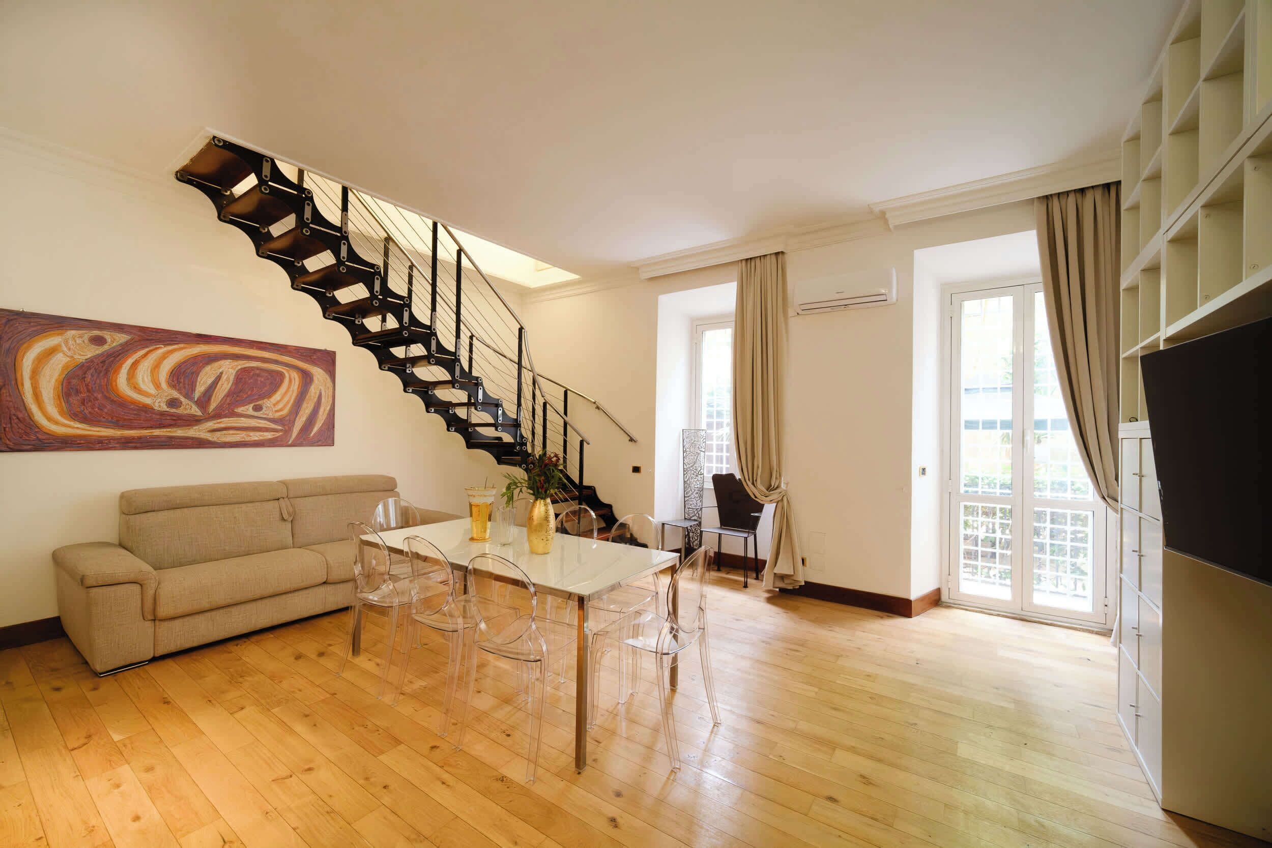 A living room with a wooden floor, a sofa, a dining table with chairs, a staircase, and a large window with curtains.