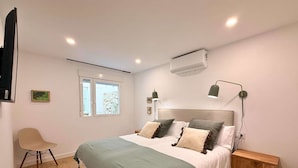 Un dormitorio con cama, mesitas de noche, una silla, una ventana y un equipo de aire acondicionado.