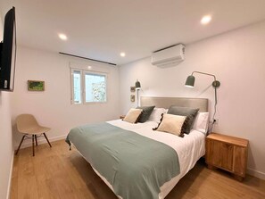 Um quarto com cama, mesas de cabeceira, uma cadeira, uma janela e um ar condicionado.