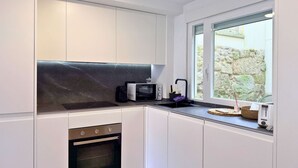 Una cocina moderna con armarios blancos, encimera negra, horno empotrado, microondas y ventana con vistas a un muro de piedra.