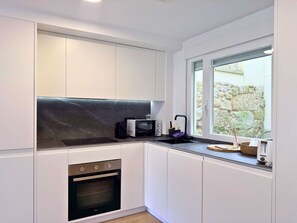 Cozinha moderna com armários brancos, bancada preta, forno embutido, micro-ondas e janela com vista para uma parede de pedra.