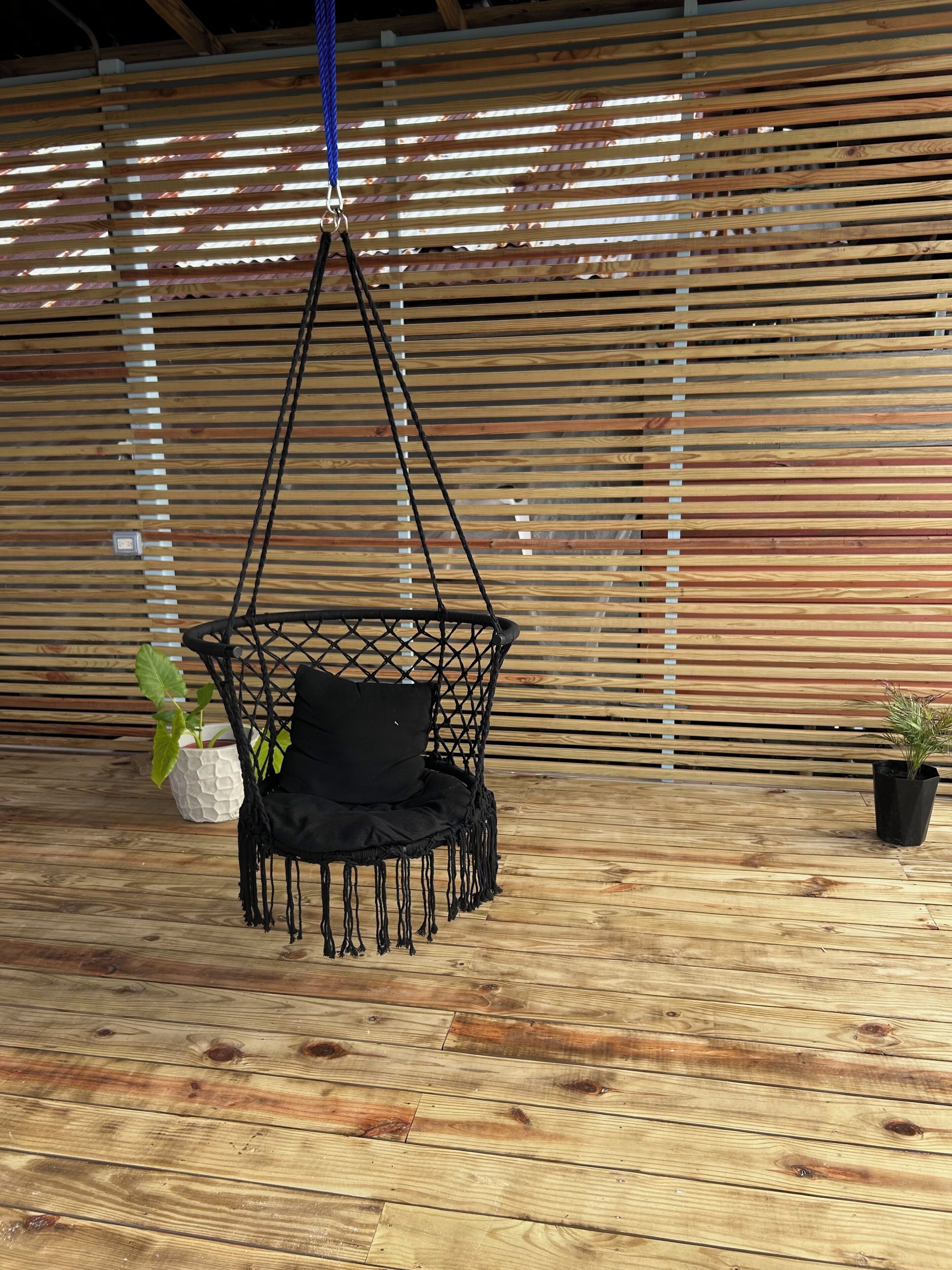 A black macrame hanging chair with a cushion, a potted plant, and a wooden floor.