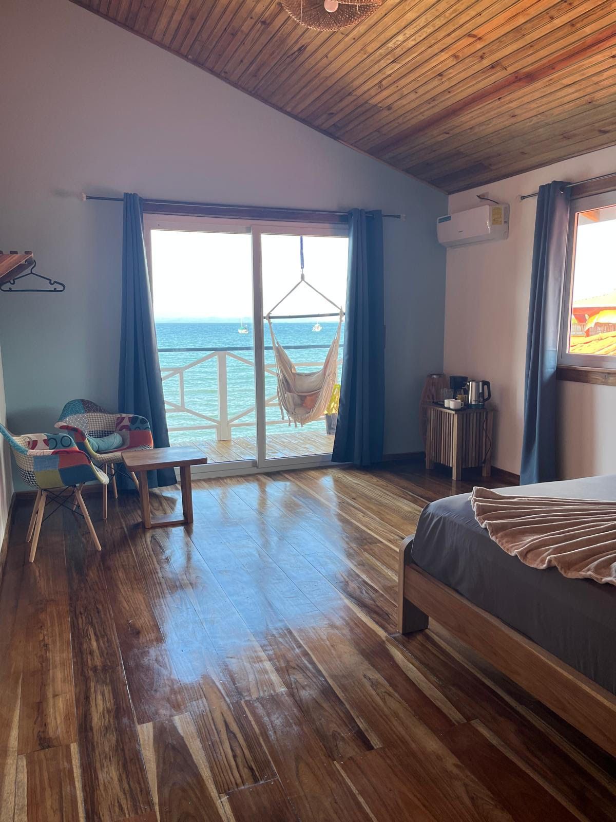 Una habitación con piso de madera, una cama con cubrecama gris, una silla colgante, una mesa con sillas y vista al mar a través de un ventanal grande.