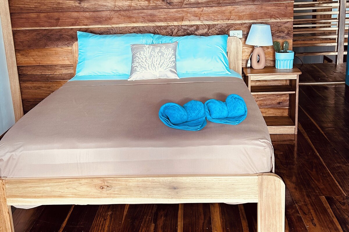 A wooden-framed bed with a beige mattress, two pillows, a blue throw, and a nightstand with a lamp and a plant.