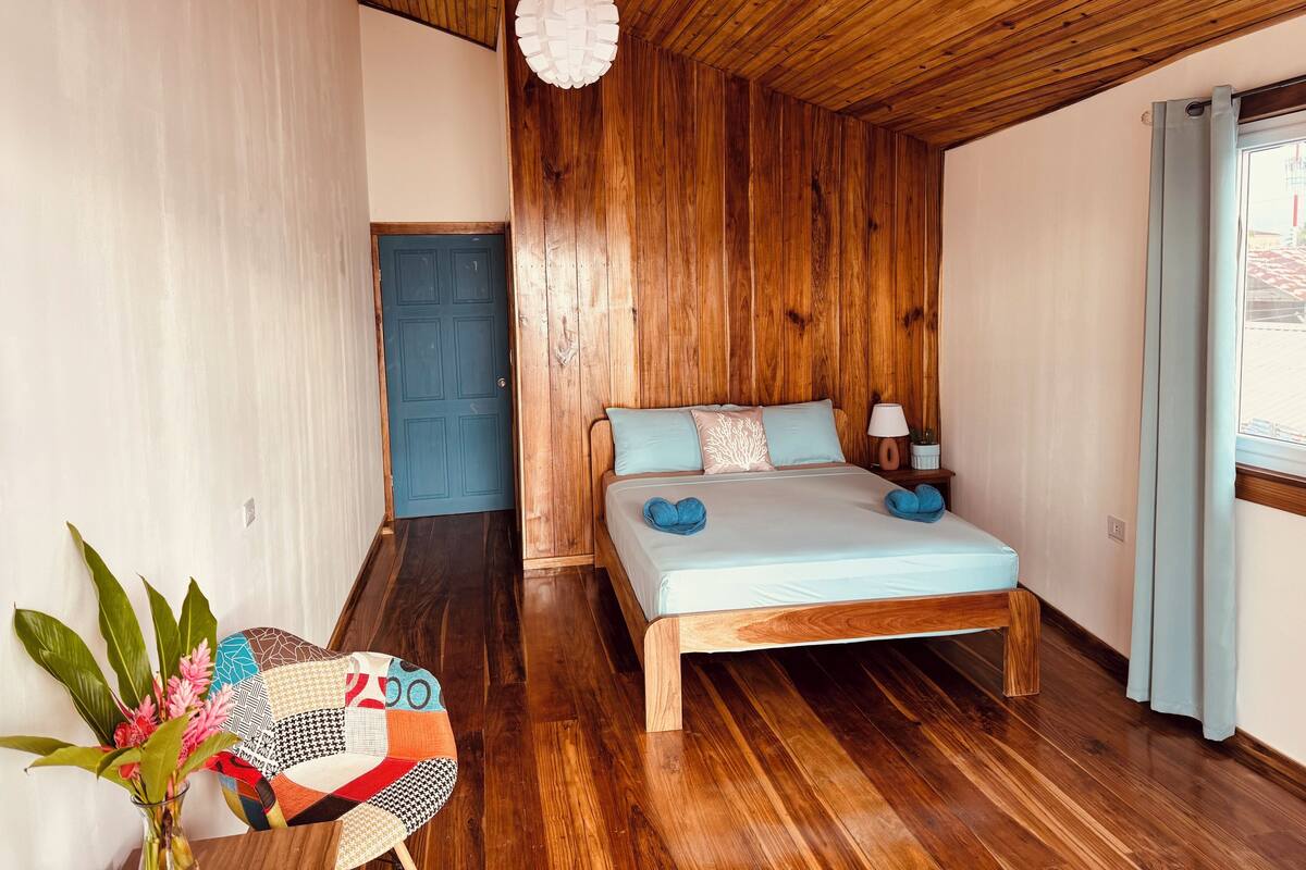 Un dormitorio con techo de madera, cama con ropa de cama azul, mesita de noche con lámpara y ventana con cortinas.