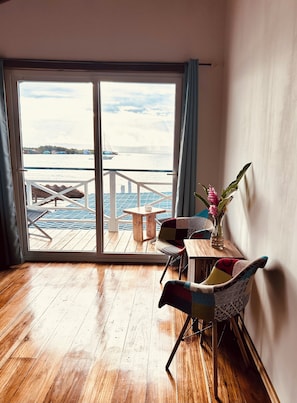 A room with a wooden floor, a view of a dock and boats, a table with chairs, and a vase with flowers.