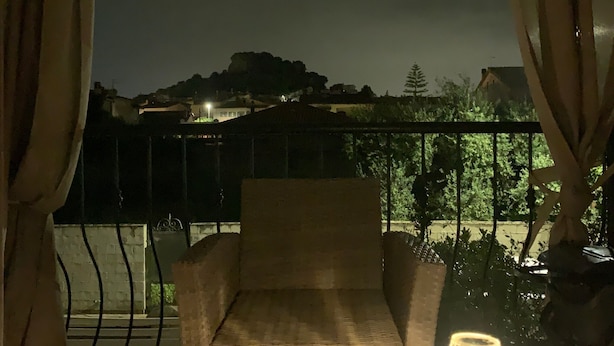 Un balcón con una silla y una mesa, una lámpara y vistas a una colina con edificios.
